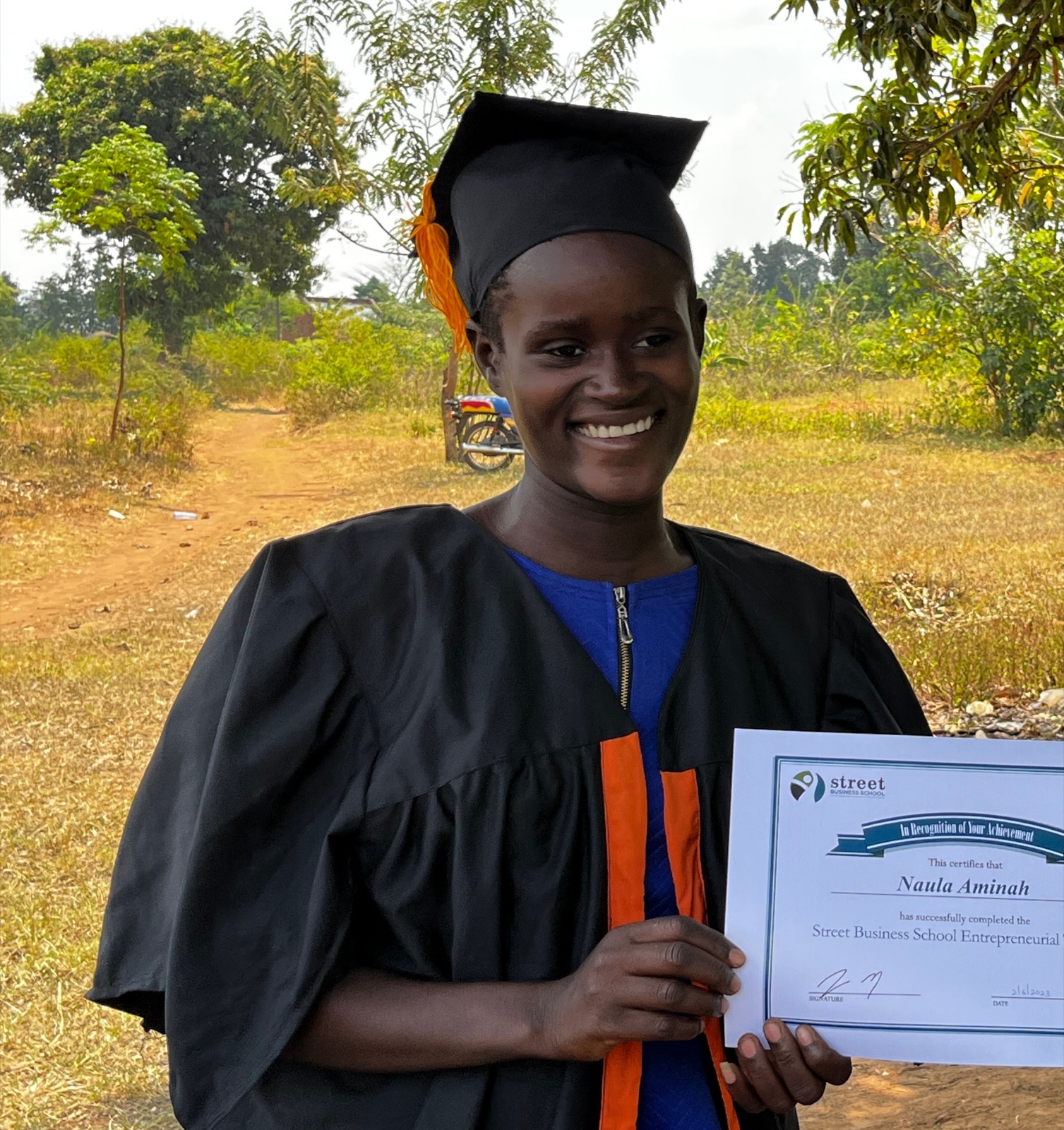 Naula Aminah smiling in graduation cap and gown, holding her Street Business School certificate alongside a GVC team member in Eastern Uganda.