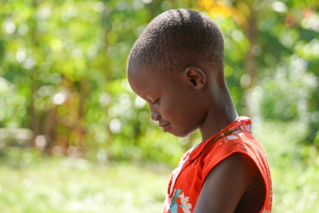 A close-up profile portrait of a young Ugandan child smiling gently against a bright, lush green background.