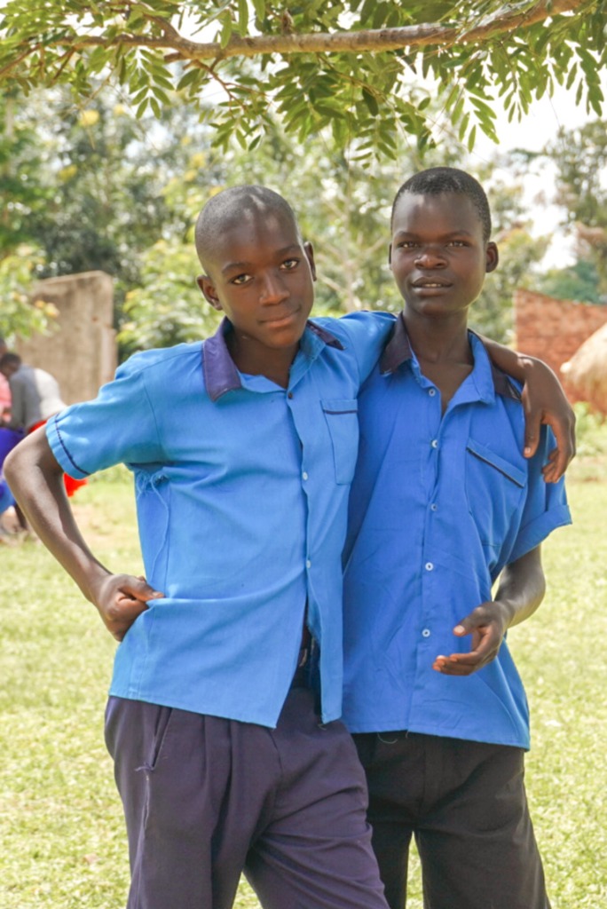 Two smiling schoolboys in bright blue uniforms standing with their arms around each other under a tree in Uganda